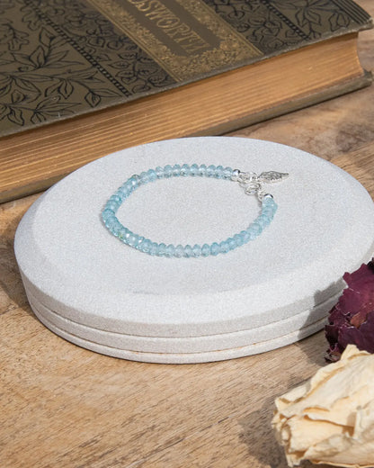 Sky blue topaz beaded gemstone bracelet with sterling silver leaf-shaped charm and spring ring clasp on light coaster on wooden table