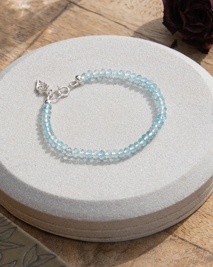 Sky blue topaz beaded gemstone bracelet with sterling silver leaf-shaped charm and spring ring clasp on light coaster on wooden table