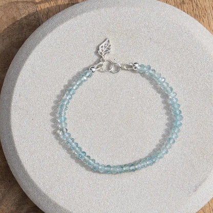 Top down photo of sky blue topaz beaded gemstone bracelet with sterling silver leaf-shaped charm and spring ring clasp on light coaster on wooden table
