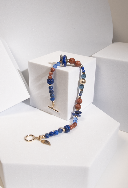 A beaded bracelet with deep blue lapis lazuli beads in round, disc, and chip shapes with sparkly burnt orange goldstone separated by 14k gold-fill accent beads and dark teal crystal beads hanging on a white cube-shaped display. A 14k gold-filled leaf-shaped charm dangles from a gold-filled toggle clasp.