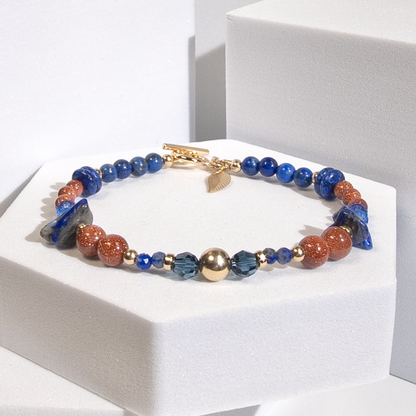 A beaded bracelet with deep blue lapis lazuli beads in round, disc, and chip shapes with sparkly burnt orange goldstone separated by 14k gold-fill accent beads and dark teal crystal beads on a white background. A 14k gold-filled leaf-shaped charm dangles from a gold-filled toggle clasp.