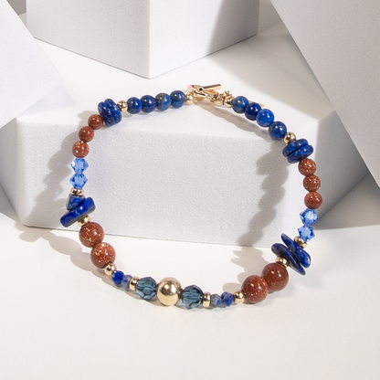 A beaded bracelet with deep blue lapis lazuli beads in round, disc, and chip shapes with sparkly burnt orange goldstone separated by 14k gold-fill accent beads and dark teal crystal beads hanging on a white cube-shaped display. A 14k gold-filled leaf-shaped charm dangles from a gold-filled toggle clasp.