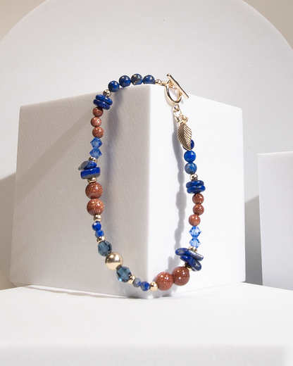 A beaded bracelet with deep blue lapis lazuli beads in round, disc, and chip shapes with sparkly burnt orange goldstone separated by 14k gold-fill accent beads and dark teal crystal beads hanging on a white cube-shaped display. A 14k gold-filled leaf-shaped charm dangles from a gold-filled toggle clasp.