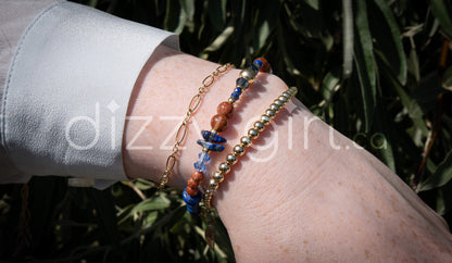 A beaded bracelet with deep blue lapis lazuli beads in round, disc, and chip shapes with sparkly burnt orange goldstone separated by 14k gold-fill accent beads and dark teal crystal beads stacked on a wrist with 14k gold-filled beaded bracelet and a 14k gold-filled paperclip chain.