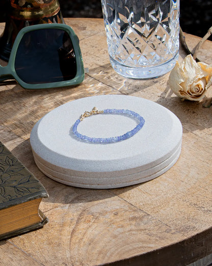 Cornflower blue faceted Tanzanite beaded bracelet in graduated size with 14k gold-filled leaf-shaped charm and spring ring clasp on light coaster on wood table