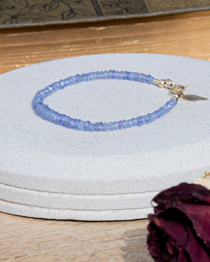 Cornflower blue faceted Tanzanite beaded bracelet in graduated size with 14k gold-filled leaf-shaped charm and spring ring clasp on light coaster on wood table