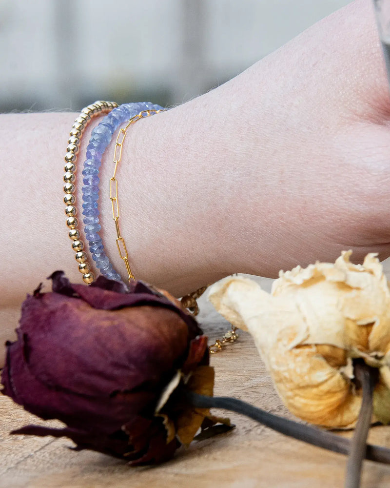 Cornflower blue faceted Tanzanite beaded bracelet stacked on wrist with 14k gold-filled beaded bracelet and 14k gold-filled paperclip chain on wooden table
