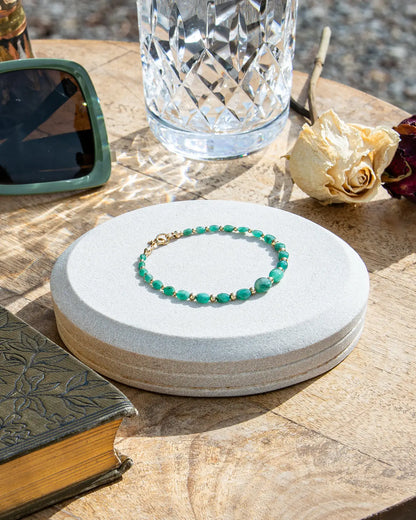 Dark green oval-shaped smooth  Zambian Emerald beaded bracelet with alternating 14k gold-filled accent beads and spring ring clasp on light coaster on a wood table