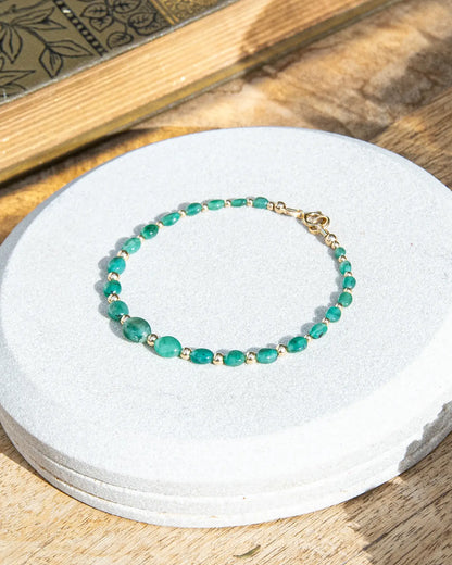 Dark green oval-shaped smooth  Zambian Emerald beaded bracelet with alternating 14k gold-filled accent beads and spring ring clasp on light coaster on a wood table