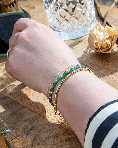 Dark green oval-shaped smooth  Zambian Emerald beaded bracelet with alternating 14k gold-filled accent beads stacked on wrist with 14k gold-filled beaded bracelet and 14k gold-filled paperclip chain on wooden table
