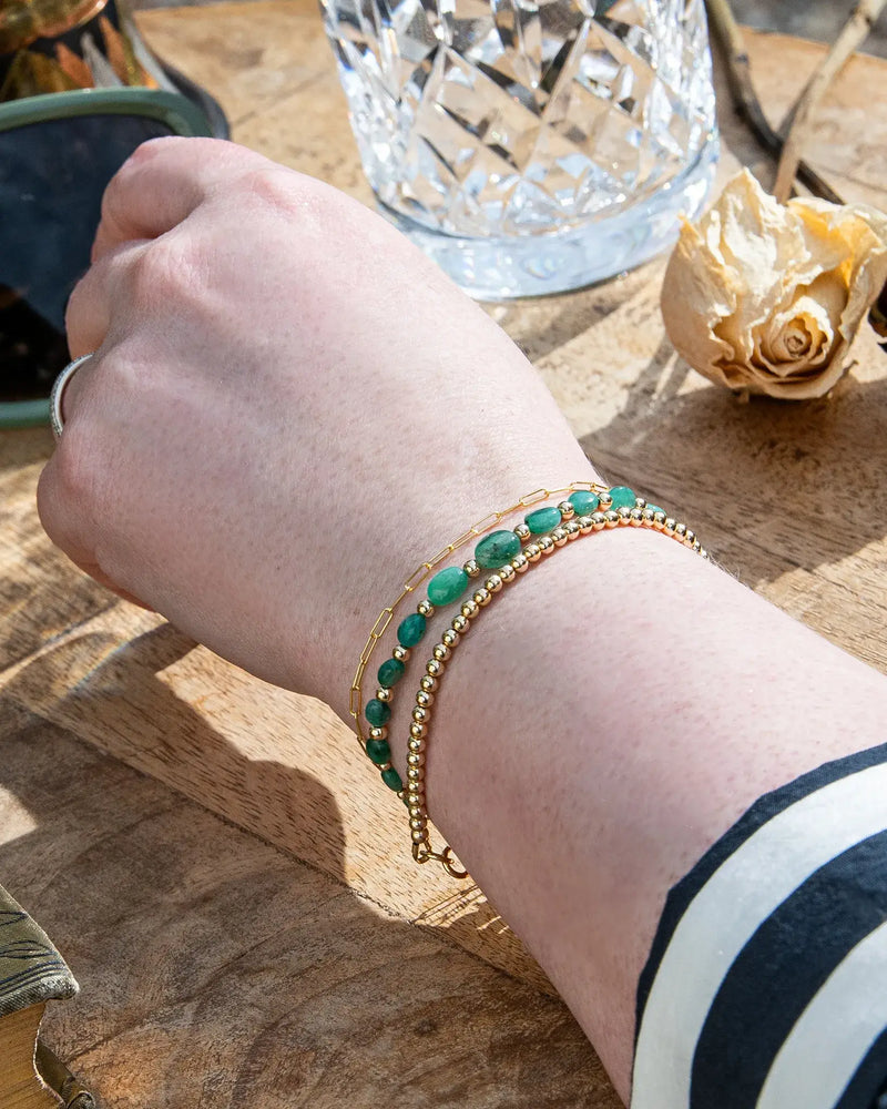Dark green oval-shaped smooth  Zambian Emerald beaded bracelet with alternating 14k gold-filled accent beads stacked on wrist with 14k gold-filled beaded bracelet and 14k gold-filled paperclip chain on wooden table